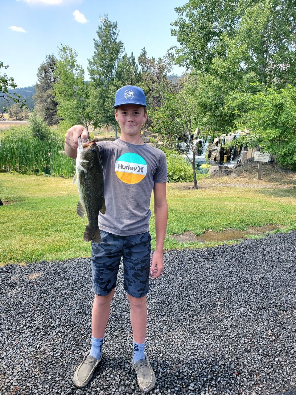 A young person standing on a gravel path holds up a largemouth bass near a grassy pond with trees and a small waterfall in the background.