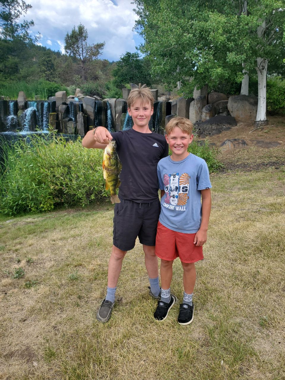 Two boys stand outdoors in front of a small waterfall, smiling as one holds up a freshly caught fish.