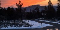 Winter sunset with orange sky over snow-covered ground and a community road sign at Ridgewater Properties in Klamath Falls, Oregon, with pine trees and snow-dusted hills in the background
